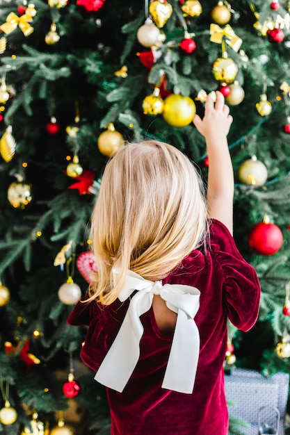 Una niña pequeña en un vestido de Borgoña con un arco se encuentra cerca del árbol de Navidad, esperando la Navidad