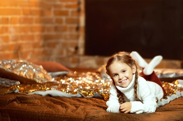 Niña pequeña con un vestido de navidad yace en la cama y sonríe Foto acogedora de la casa Confort navideño en casa