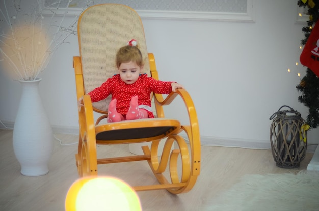 Niña pequeña con vestido rojo jugando en la habitación con adornos navideños en una mecedora