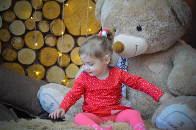 Niña pequeña con vestido rojo jugando en la habitación con adornos navideños y oso grande