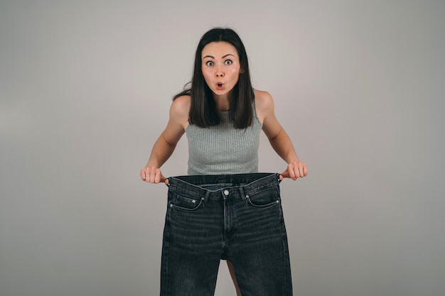 La niña perdió mucho peso. La niña sostiene jeans muy grandes. La mujer se sorprende de cómo perdió peso. Problemas de la anorexia femenina.