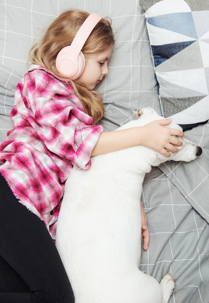 Niña y perro acostados en el sofá con auriculares, escuchando música con su teléfono inteligente.