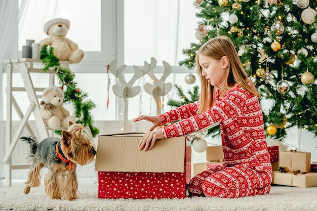 Niña con perro desembala caja de regalo roja de Navidad en casa. Niño celebrando el año nuevo con regalos y mascota perrito