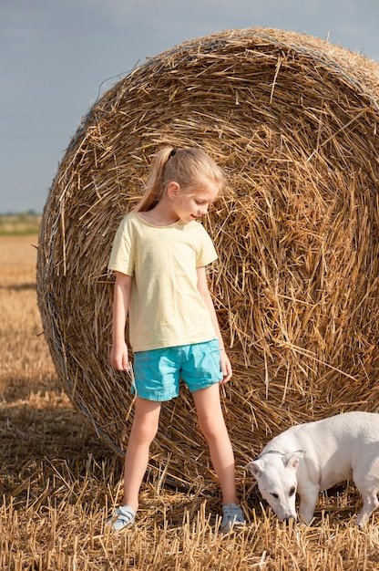 Niña y perro divirtiéndose en un campo de trigo en un día de verano. Niño jugando en el campo de balas de heno durante la época de la cosecha.