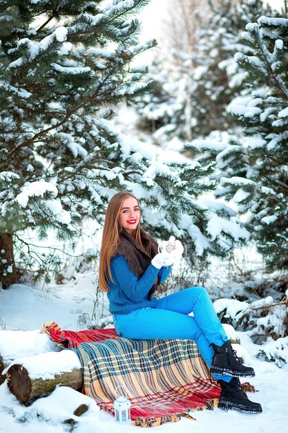 Una niña en un picnic de invierno en un bosque de coníferas cubierto de nieve Ropa cálida y elegante Taza con té en las manos