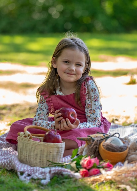 Niña en un picnic. Sostiene una manzana roja en sus manos y se para junto a una canasta de frutas.