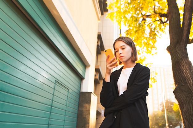 Niña de pie en la calle en el fondo de un árbol en la luz del sol y la arquitectura y usando un teléfono inteligente