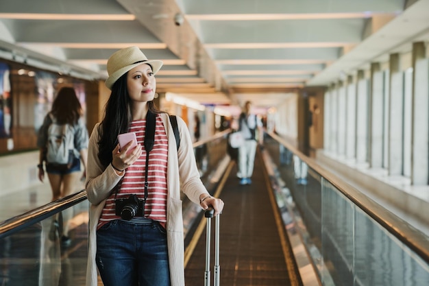 niña de pie en una larga escalera mecánica horizontal en la terminal del aeropuerto internacional mano sosteniendo el horario de control del teléfono celular del horario de vuelo del avión. mujer asiática con equipaje en la pasarela móvil.