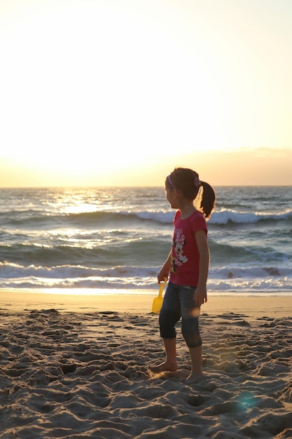 Foto niña de pie en la playa durante la puesta de sol