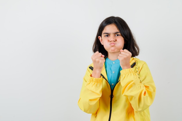 Niña de pie en pose de lucha mientras sopla las mejillas en camisa, chaqueta y parece confiada. vista frontal.