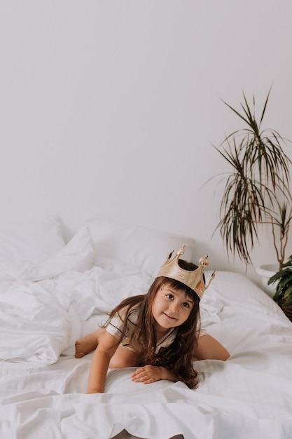 niña en pijama dorado y una corona en una cama en casa con sábanas blancas. mañana del pequeño