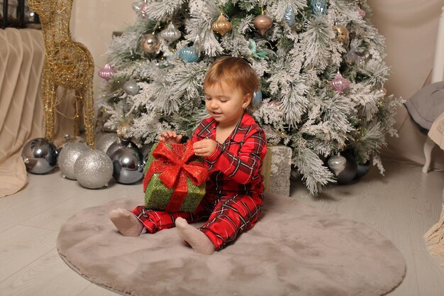 una niña en pijama de Navidad está sentada en el árbol de Navidad con una caja de regalo en sus manos