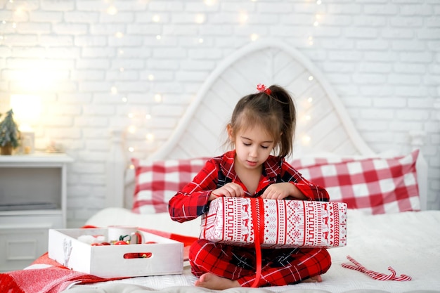 Una niña en pijama rojo a cuadros desenvuelve un regalo de Navidad mientras está sentada en la cama