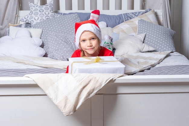 Una niña en pijama rojo y un gorro de Papá Noel sostiene una gran caja de regalo en la cama en la guardería Regalos de Navidad y Año Nuevo