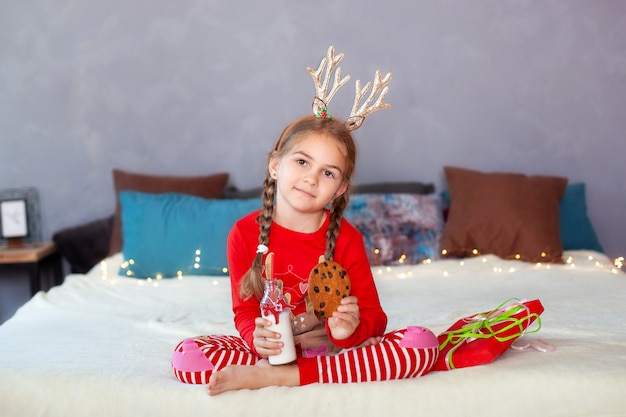 niña en pijama rojo se sienta con leche y galletas en la víspera de Navidad y espera a Santa Claus. El niño come galletas con leche en casa. Niña vestida como un cuerno de ciervo. Feliz navidad año nuevo