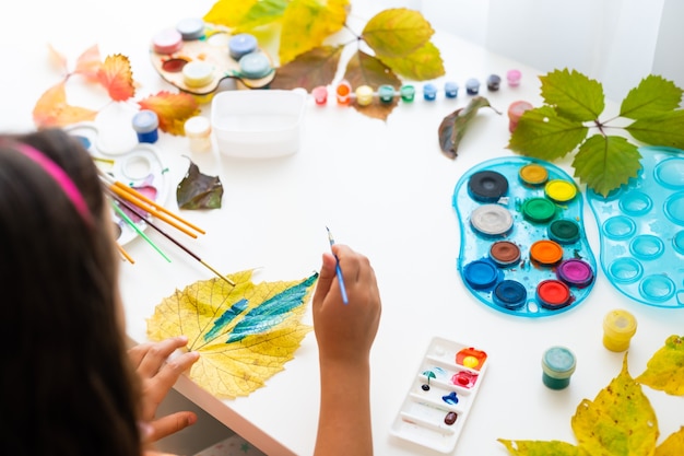 niña pinta hojas con pincel en casa, arte de otoño.