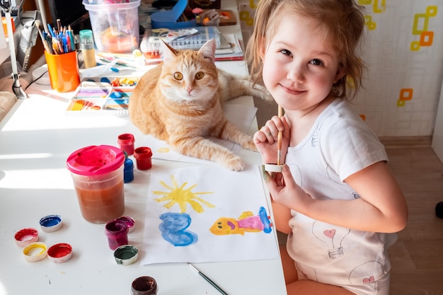 Foto una niña pinta el sol y su madre con acuarelas un gato pelirrojo yace junto a la mesa.