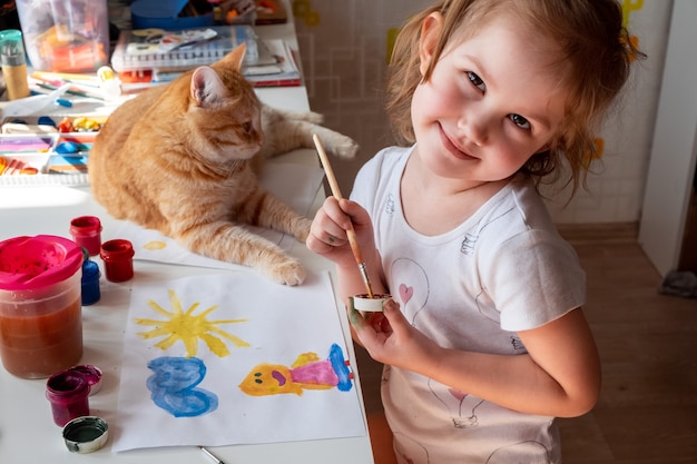 Una niña pinta el sol y su madre con acuarelas un gato pelirrojo yace junto a la mesa.