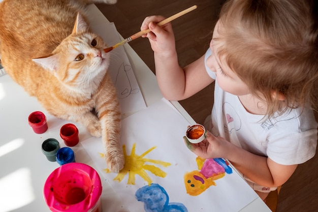 Una niña pinta el sol y su madre con acuarelas un gato pelirrojo yace junto a la mesa.