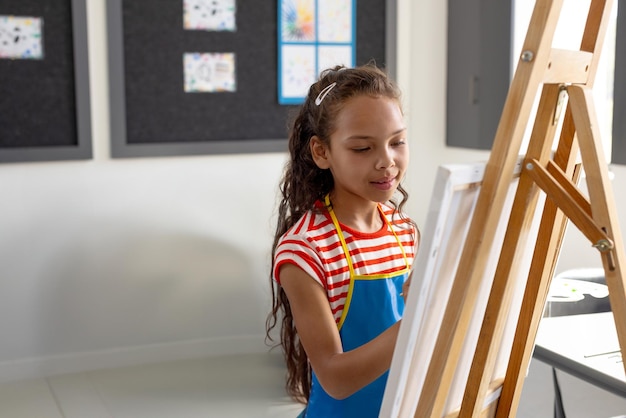 Niña pintando en lienzo en caballete en la clase de arte con pincel y dibujos fijados