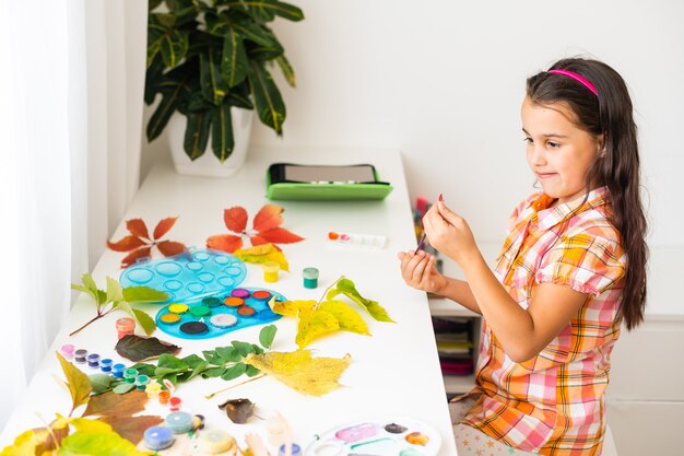 Niña pintando sobre hojas amarillas otoñales con gouache, artes infantiles, creatividad infantil, arte otoñal.