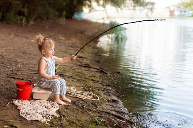 Niña en una playa de arena cerca del río sostiene una caña de pescar y atrapa peces