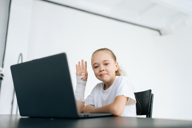 Niña positiva con la mano rota envuelta en una venda de yeso blanco que muestra un gesto de hola mirando a la cámara sentada en el escritorio con una laptop en la sala de luz