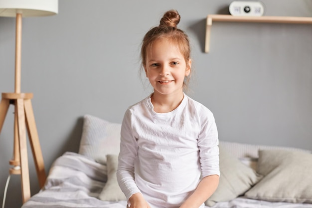 Niña positiva sentada en la cama niño sonriente con peinado de moño con ropa blanca para el hogar mirando a la cámara con expresión optimista lindo niño emocional