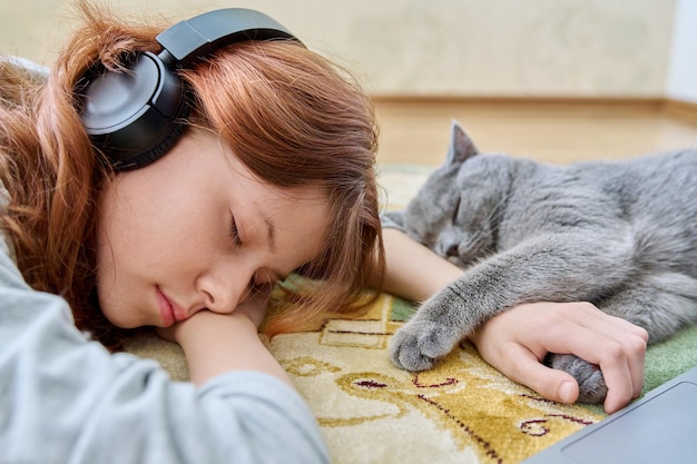 Niña preadolescente en auriculares durmiendo con gato en el suelo en casa El joven propietario y la mascota británica gris duermen juntos Los animales aman la amistad concepto de estilo de vida de los niños