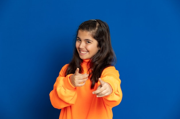 Niña preadolescente con jersey amarillo gesticulando sobre pared azul