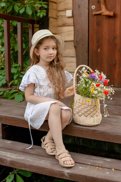 Niña preadolescente soñadora con ropa de verano y sombrero de paja sosteniendo un montón de flores silvestres coloridas en una canasta de mimbre mientras se sienta en la puerta de la casa de campo disfrutando el fin de semana de verano