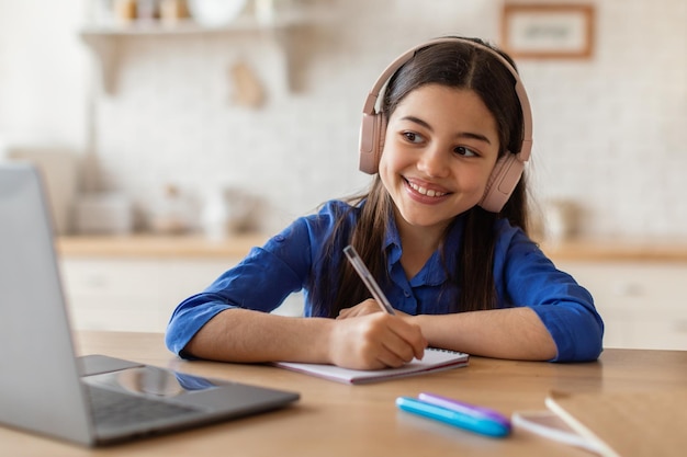 Niña preadolescente sonriente en auriculares aprendiendo con laptop en casa