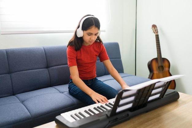 Niña preadolescente tocando el teclado en su sala de estar mientras escucha una canción con auriculares. Chica hispana practicando el piano con una partitura