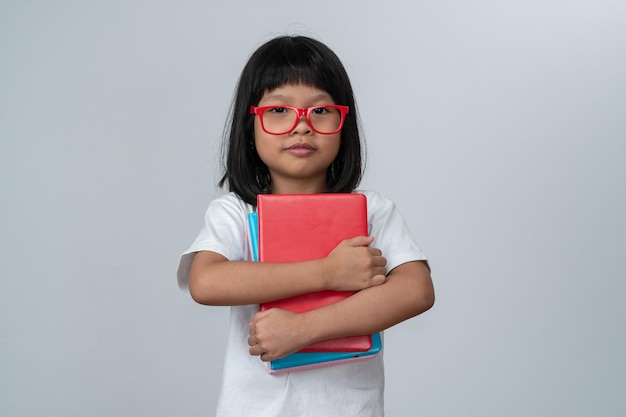 Niña preescolar asiática feliz con gafas rojas sosteniendo libros y mochila roja