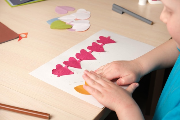 Foto niña preescolar con camiseta azul hace apliques sentados en la mesa, corazones cortados de bougue de colores para la tarjeta para el día de san valentín, creatividad infantil, dermatitis atópica en las manos de un niño