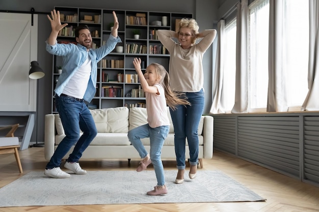 Una niña preescolar encantada bailando con un padre joven y una abuela madura en la sala de estar felices tres generaciones familiares se divierten participando en una divertida actividad de movimiento en casa juntos el fin de semana