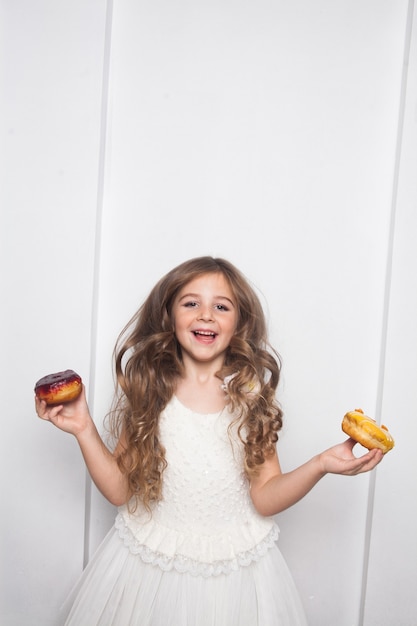 Niña preescolar linda feliz está comiendo donuts coloridos sobre fondo blanco aislado.