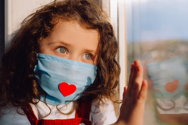 Foto niña preescolar en mascarilla médica estéril con corazón rojo, sentado en el alféizar de la ventana en casa, de la mano sobre el vidrio, mirando fuera de la ventana borrosa. quedarse en casa y el concepto de cuarentena