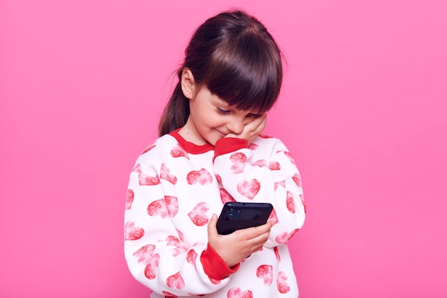 Niña preescolar pequeña de cabello oscuro con cabello oscuro y coleta sosteniendo el teléfono inteligente en las manos, mirando su pantalla, tocando su rostro y sonriendo, posando aislada sobre la pared rosa.