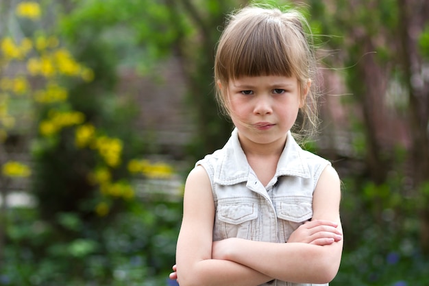 La niña preescolar rubia cambiante bastante divertida en el vestido sin mangas blanco mira en la cámara sintiéndose enojado e insatisfecho en el fondo borroso del verano. Concepto de berrinche de los niños.