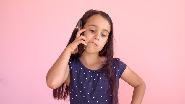 Niña preescolar con un vestido sosteniendo un teléfono móvil, aislada sobre rosa