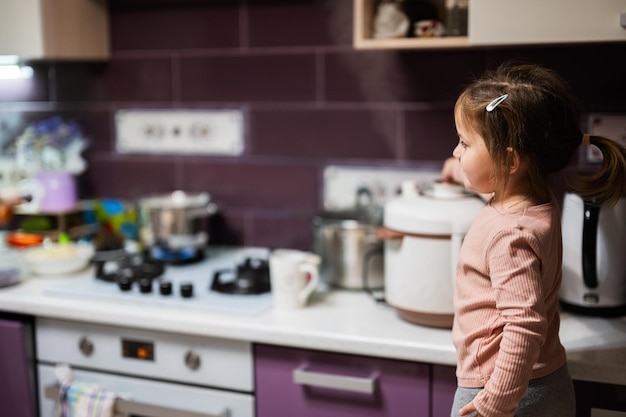 La niña prepara la cena en la cocina de casa con una olla de cocción lenta y libera vapor del vapor