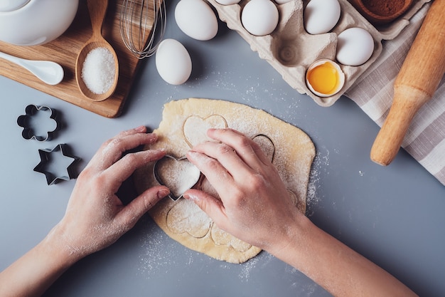 Niña prepara galletas en forma de corazón, composición laica plana sobre un fondo gris. Cortadores de galletas y masa en manos de mujeres. Concepto de comida para el día de San Valentín, día del padre, día de la madre.