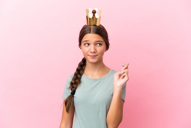 Niña princesa caucásica aislada sobre fondo rosa con los dedos cruzando y deseando lo mejor