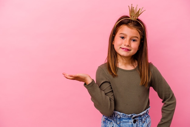 Niña princesa con corona aislada en rosa mostrando un espacio de copia en una palma y sosteniendo otra mano en la cintura.