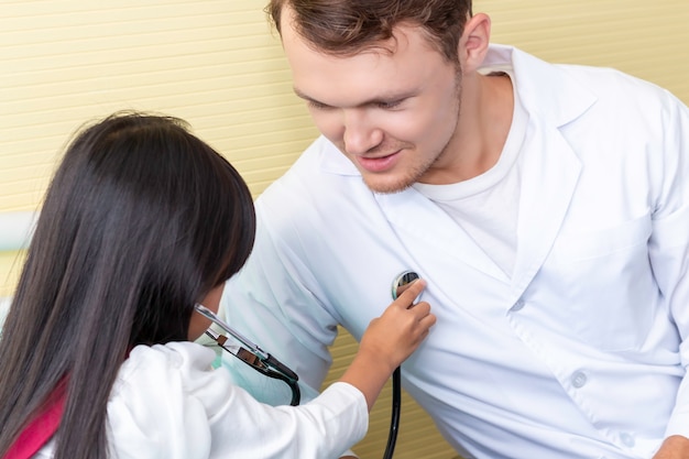 Foto niña que juega y que examina al hombre del pediatra que usa un estetoscopio en hospital del dormitorio.