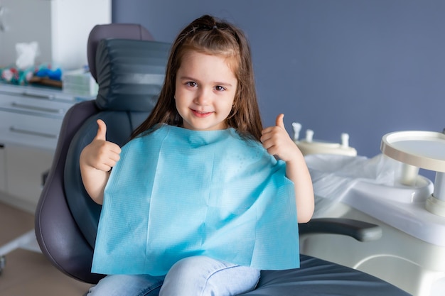 Una niña que lleva un delantal azul se sienta en una silla de dentista y da un pulgar hacia arriba.