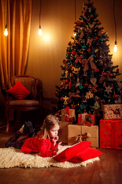 Niña que miente en alfombra con los regalos alrededor usando la tableta en la almohada roja.