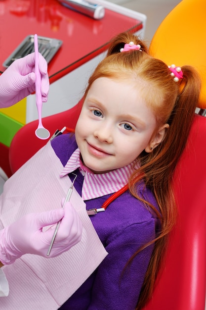 Niña que sonríe en silla dental roja. El dentista examina los dientes del paciente del niño.