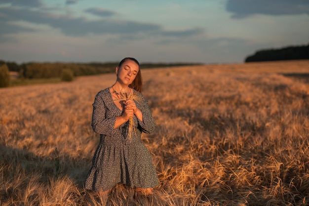 Una niña con un ramo de espigas de trigo se para en un campo de trigo al atardecer